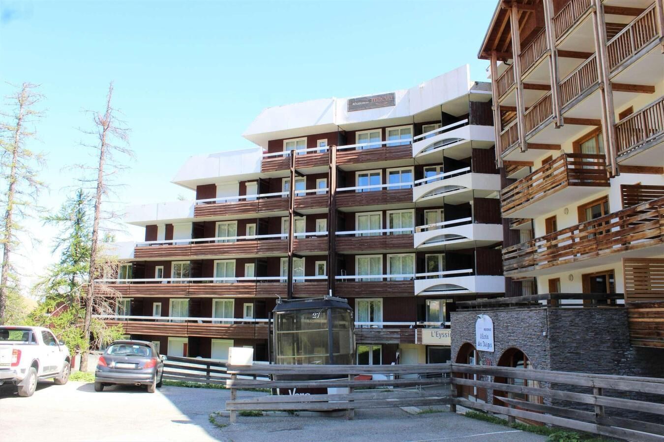 A multi-story hotel with balconies, a stone facade, and a parking area with cars.