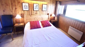 Una habitación con paneles de madera, una cama, dos mesitas de noche, una silla y cuadros enmarcados en la pared.