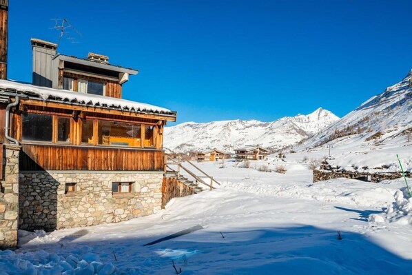 A wooden chalet with stone walls, a wooden deck, and a view of snow-covered mountains.