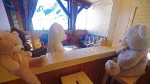 Una zona de bar de madera con dos grandes osos de peluche sentados en la barra, un taburete de madera y varios vasos de vino sobre la encimera.