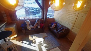 Interior acogedor de una cabaña con dos sofás, una mesa de comedor y vistas a un paisaje nevado a través de amplias ventanas.