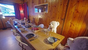 Una mesa de comedor de madera dispuesta para seis personas, con sillas, un oso de peluche y una televisión mostrando un paisaje urbano.