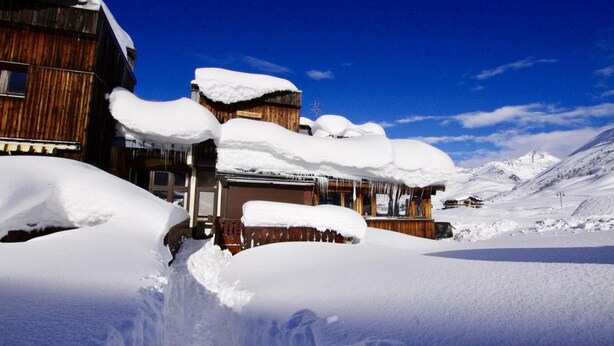 Un chalet de madera cubierto de nieve, con un cielo azul despejado y montañas nevadas al fondo.