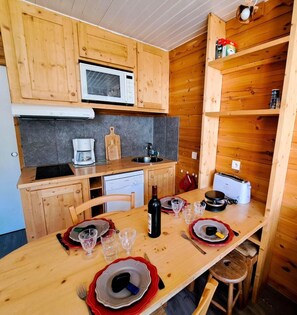 Een kleine keuken met houten kastjes, een magnetron, een koffiezetapparaat en een eettafel voor twee personen, gedekt met borden, glazen en bestek.