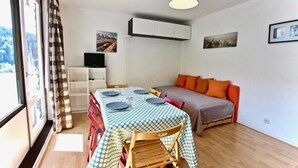 A compact room with a dining table set for four, a sofa with orange cushions, a TV on a shelf, and a cityscape view through a window. - 150m from ski slopes, 3rd floor, balcony, tv, 25m², Chamrousse (Chamrousse)