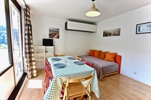 Habitación pequeña con una mesa de comedor para cuatro personas, un sofá con cojines naranja, un televisor en una estantería y vistas a la ciudad a través de una ventana.