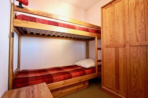 A bunk bed with a red checkered bedspread and a white pillow. There is a wooden wardrobe to the right and a small wooden table in front of the bed.