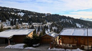 Un albergue de montaña con fachada de madera, zona de estacionamiento cubierta y vistas a montañas nevadas y laderas boscosas.