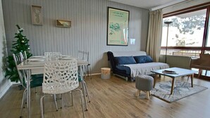 Una sala de estar con una mesa de comedor, sillas, un sofá y una mesa de centro. Hay un pequeño árbol de Navidad y un cartel en la pared.