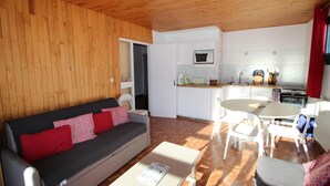 Un espace de vie compact doté d’un plafond en bois, d’un canapé avec des coussins rouges et blancs, d’une table à manger avec des chaises, d’une kitchenette équipée d’un four à micro-ondes et d’une cuisinière, et d’une petite fenêtre.