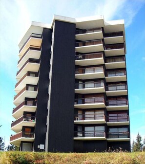 Un immeuble résidentiel à plusieurs étages, doté de balcons, avec une section verticale centrale de couleur sombre, et un ciel dégagé en arrière-plan.
