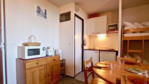 Cocina compacta con microondas, tostadora y cafetera sobre una encimera de madera. En primer plano, una mesa de comedor con sillas, y al fondo se ve una cama nido.