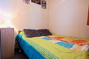 Quarto pequeno com cama de solteiro, cabeceira de madeira, colcha colorida e luminária na parede.