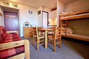 A compact room with a bunk bed, a dining table with chairs, a microwave, and a red plaid sofa.