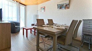 Zona de menjador amb taula de fusta preparada per a quatre, envoltada de cadires, una panxa vermella i un escriptori amb un ordinador.