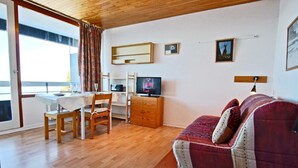 Een gezellige woonkamer met een houten vloer, een rode bank, een televisie, een eettafel met stoelen en een raam met gordijnen.
