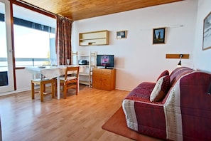 A cozy living room with a wooden floor, a red sofa, a TV, a dining table with chairs, and a window with curtains. - 100m from ski slopes, 2nd floor, balcony, 27m², Chamrousse (Chamrousse)