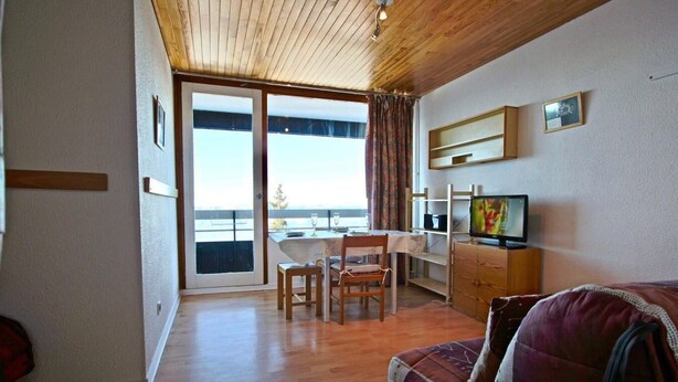 Een kamer met een houten plafond, een eethoek met een tafel en stoelen, een televisie op een standaard en uitzicht door een glazen deur.