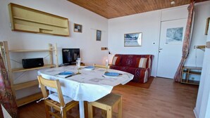 Een eetgedeelte met een ronde tafel gedekt voor vier personen, houten stoelen, een televisie op een plank, en een rode, gestreepte bank op de achtergrond.