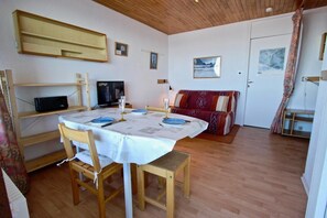 A dining area with a round table set for four, wooden chairs, a television on a shelf, and a red patterned sofa in the background. - 100m from ski slopes, 2nd floor, balcony, 27m², Chamrousse (Chamrousse)