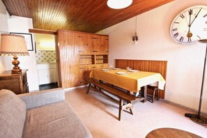 Un coin repas avec une table en bois dressée pour deux personnes, un banc, un canapé et une horloge murale.