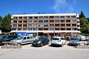 Un hôtel à plusieurs étages, doté d’une rangée de balcons, un parking où sont garées plusieurs voitures, et diverses devantures de magasins au premier plan.