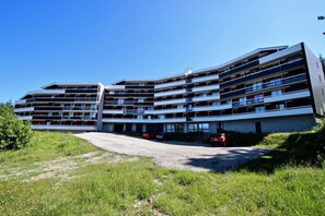 A multi-story building with balconies, a parking area, and a grassy foreground.