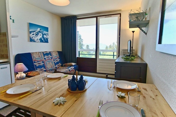 A dining area with a wooden table set for two, a sofa with a colorful quilt, a window with a view of greenery, and a small cabinet with a lamp.