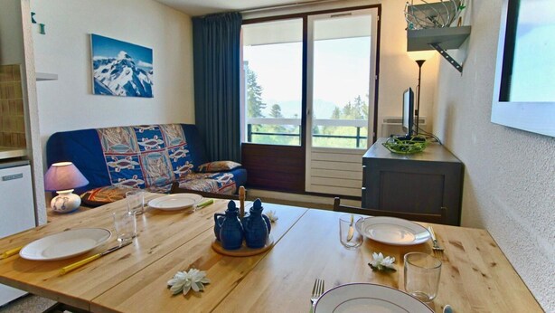 A dining area with a wooden table set for two, a sofa with a colorful quilt, a painting of a mountain, and a view through the window. - 100m from ski slopes, 3rd floor, tv, ski locker, 21m², Chamrousse (Chamrousse)