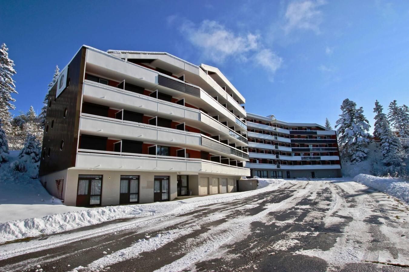 Hotel de varios pisos con balcones, zona de estacionamiento y alrededores nevados.