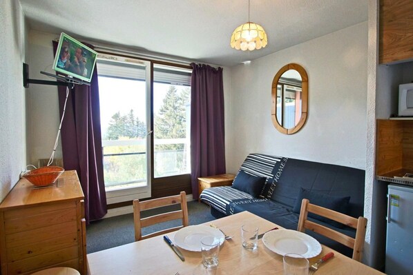 A compact room with a dining table set for two, a sofa, a television on a stand, and a large window with curtains.