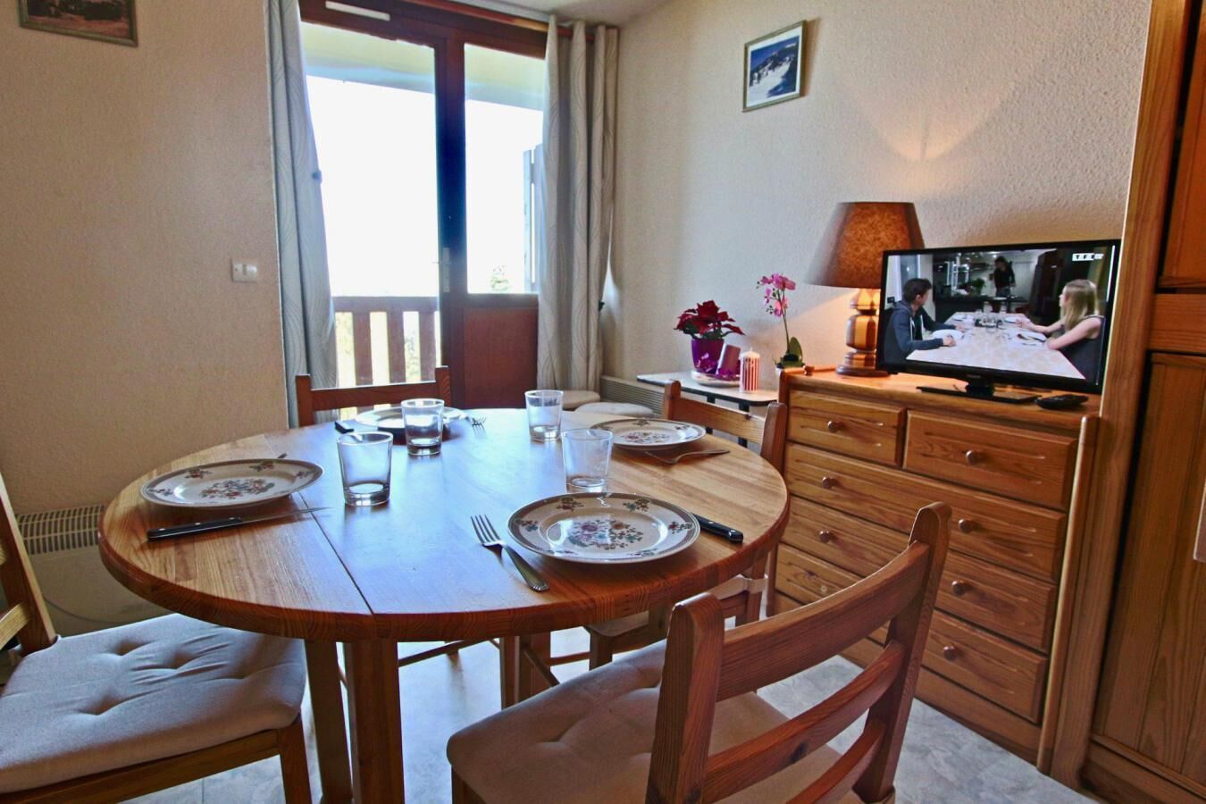Un comedor con una mesa redonda de madera dispuesta para cuatro personas, sillas, un televisor sobre un mueble de madera y una ventana con cortinas.