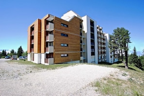 A multi-story building with a mix of wooden and white exterior panels, balconies, and a gravel driveway.