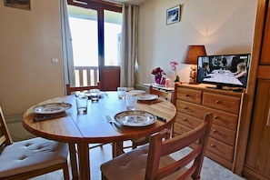 A dining area with a round table set for four, chairs, a television on a wooden cabinet, and a window with curtains.
