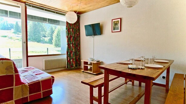Une pièce avec une table en bois dressée pour le repas, un canapé, un téléviseur accroché au mur et une vue sur l’extérieur grâce à une grande fenêtre.