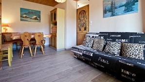 Un salon chaleureux avec un canapé orné de noms de villes, un coin repas avec des chaises en bois et un tableau représentant un paysage enneigé.