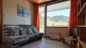 Un salon avec un canapé orné de noms de villes, un tableau représentant des arbres, un radiateur et un téléviseur posé sur un meuble en bois.