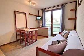 Een knus ingerichte kamer met een houten eettafel en stoelen, een bank met kussens, een televisie op een meubel en een raam met gordijnen.