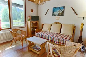 Ein gemütliches Zimmer mit einer Couch, zwei Korbsesseln, einem kleinen Tisch, einem Fernseher und einem Fenster mit Blick auf Bäume.