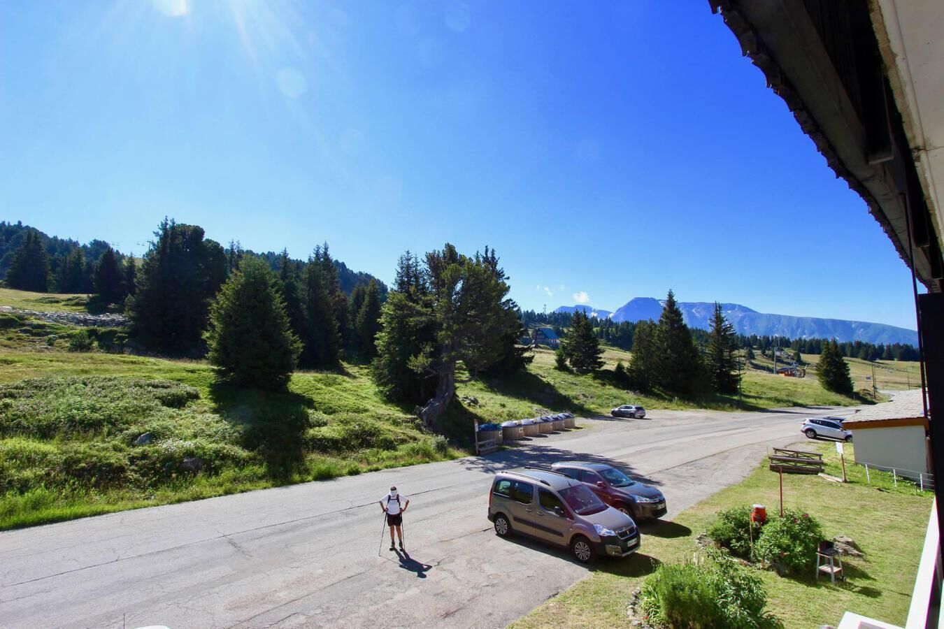 A parking area with cars and a person skiing, surrounded by greenery and mountains in the background.