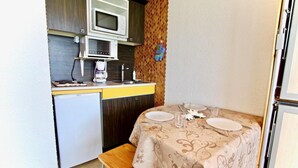 Une petite cuisine équipée d’un four à micro-ondes, d’une machine à café et d’un petit réfrigérateur. Une table à manger avec une nappe à motifs floraux et un banc sont installés dans un coin.