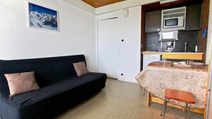 A compact living space with a black sofa, a small dining table with a patterned tablecloth, and a kitchenette area with a microwave and a coffee maker. - 100m from ski slopes, Ground floor, 19m², Chamrousse (Chamrousse)