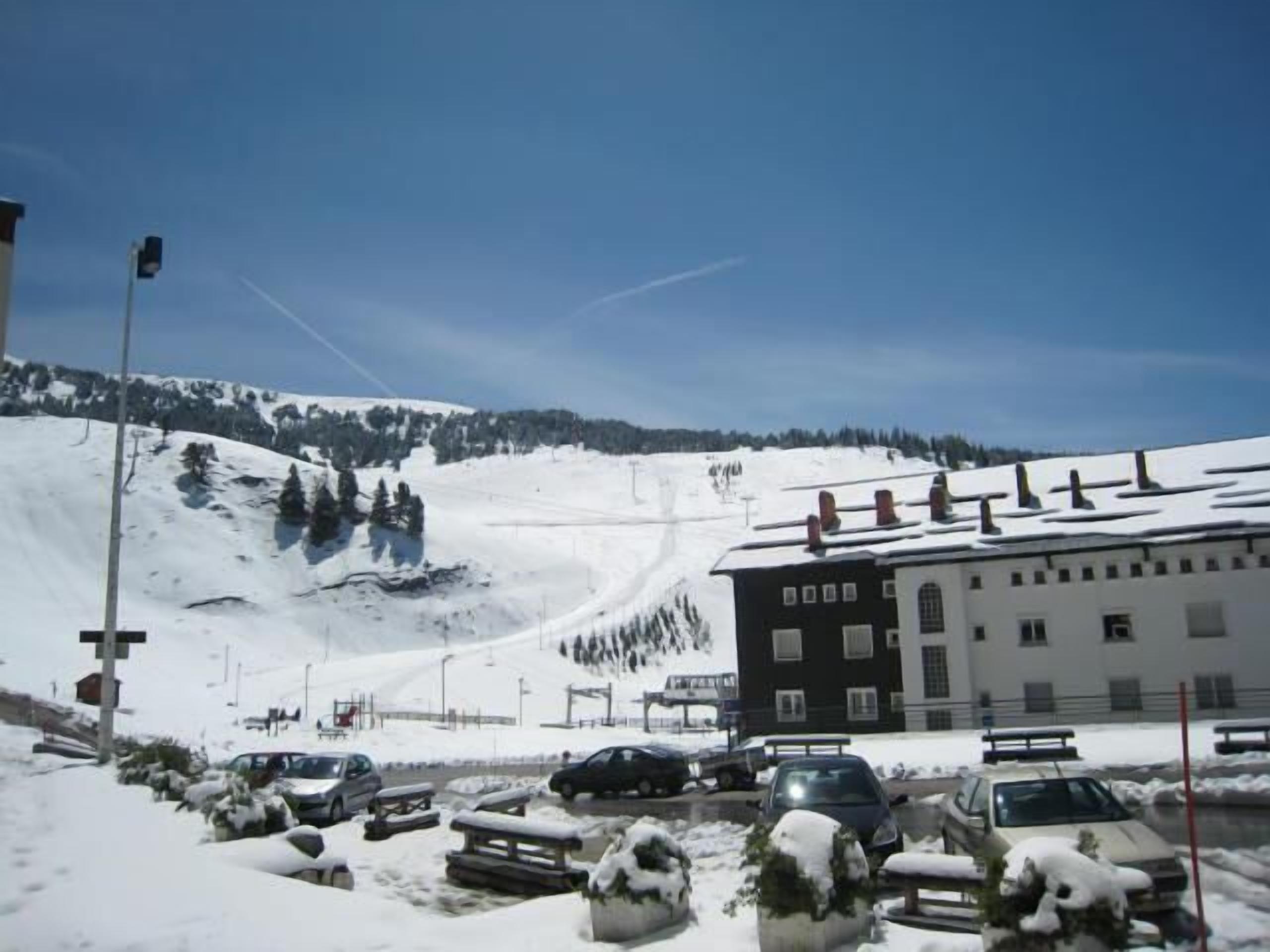 A snow-covered parking lot with several cars, a building with multiple windows, and a ski slope in the background.