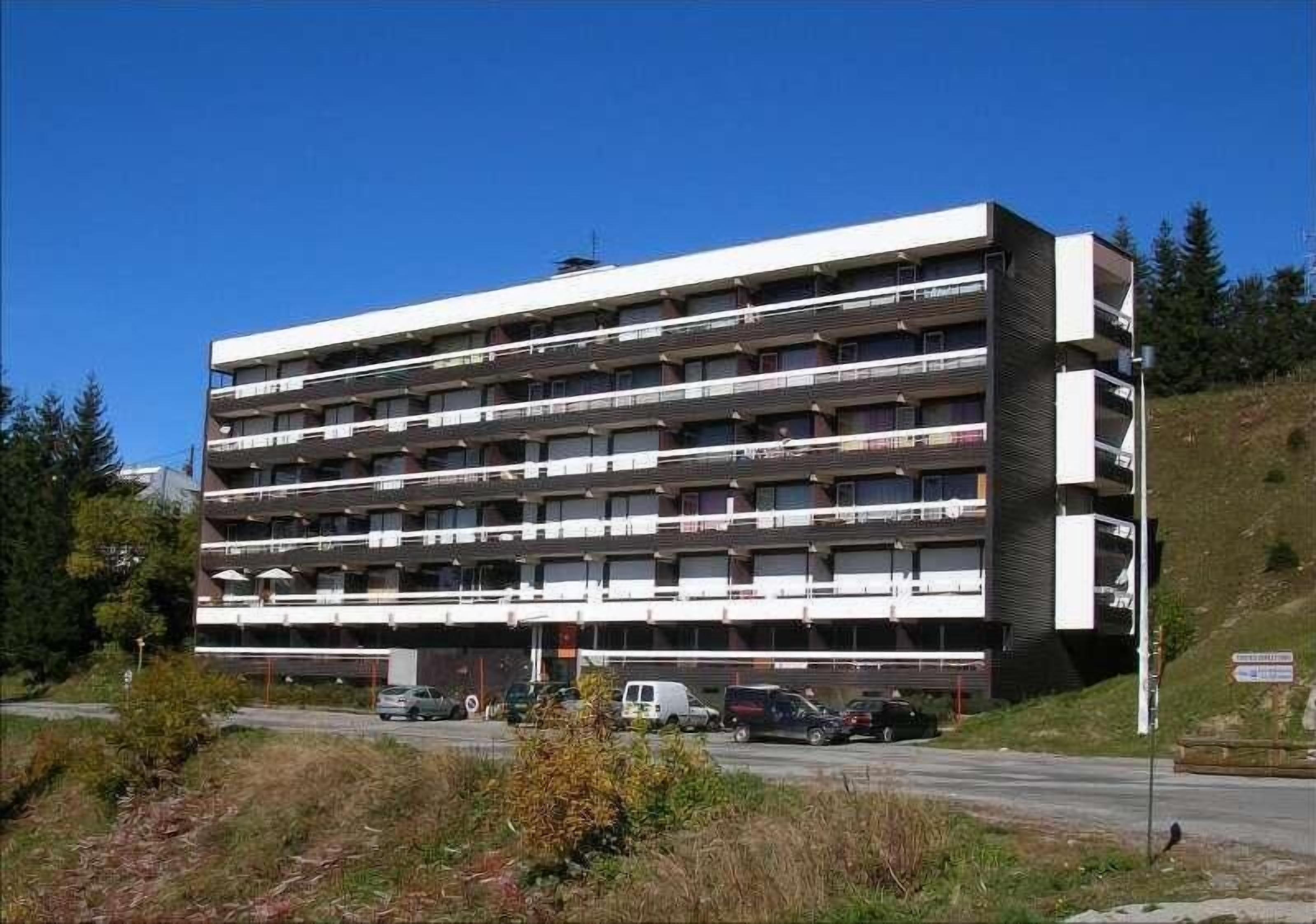 Un edificio de varios pisos con balcones, situado cerca de una calle con coches estacionados.