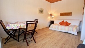 Une pièce avec un plancher en bois, un lit recouvert d’une couverture à rayures colorées et agrémenté d’oreillers orange, une table à manger avec une nappe à motifs floraux et deux chaises, ainsi qu’une lampe fixée au mur.
