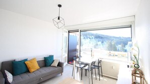 A modern living room with a grey sofa, colorful cushions, a dining table with chairs, and a large window offering a view of a forested area. - 150m from ski slopes, 2nd floor, balcony, tv, 30m², Chamrousse (Chamrousse)