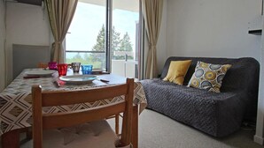 Un espace de vie chaleureux avec une table à manger pour deux personnes, un canapé agrémenté de coussins à motifs et une grande fenêtre avec des rideaux.