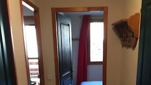 A narrow hallway leading to a bedroom with a bed, a window with red curtains, and a door on the left.