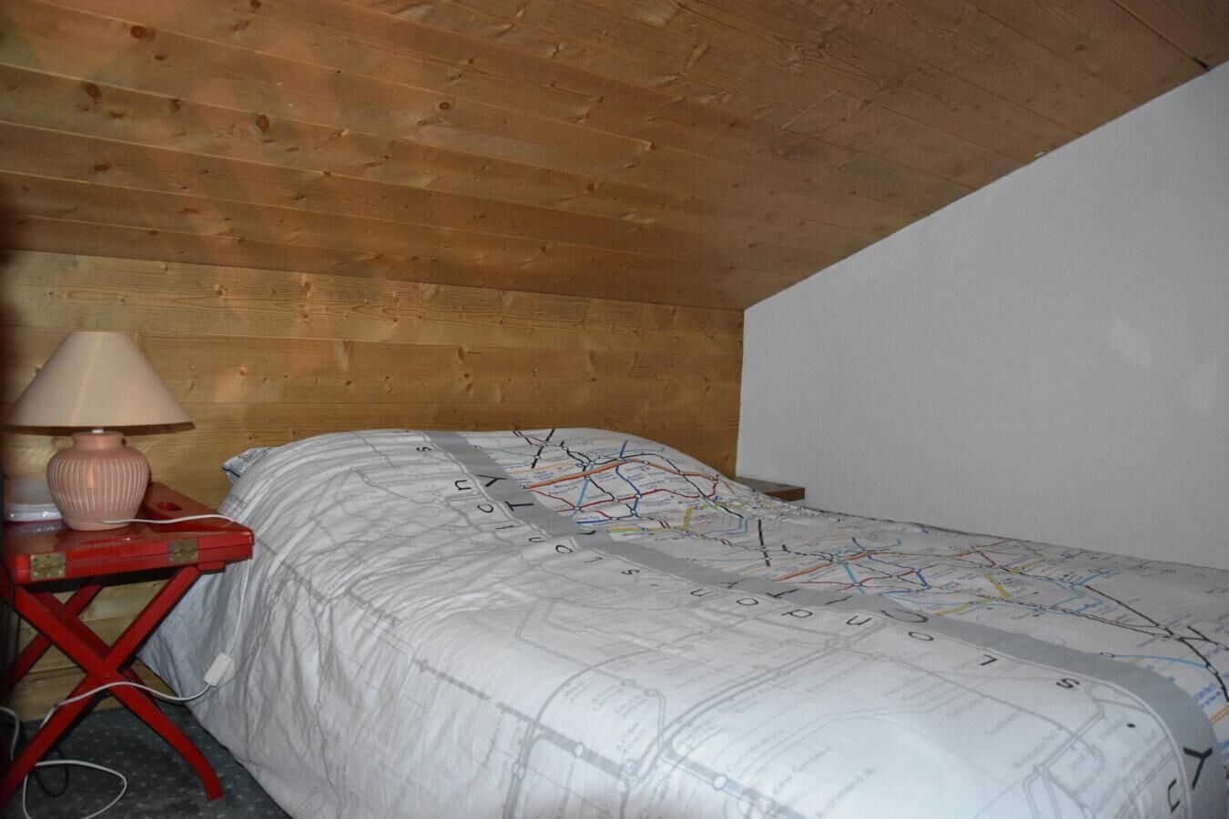Ein Schlafzimmer mit Holzboden, einem Einzelbett mit Bettwäsche mit Weltkarte, einem roten Nachttisch mit Lampe und einer weißen Wand.