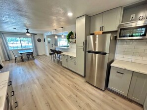 A modern kitchen with stainless steel appliances, light wood flooring, and a dining area with bar stools. - Modern Chic 4-Bedroom Home with Wood Stove Fireplace (Flagstaff)